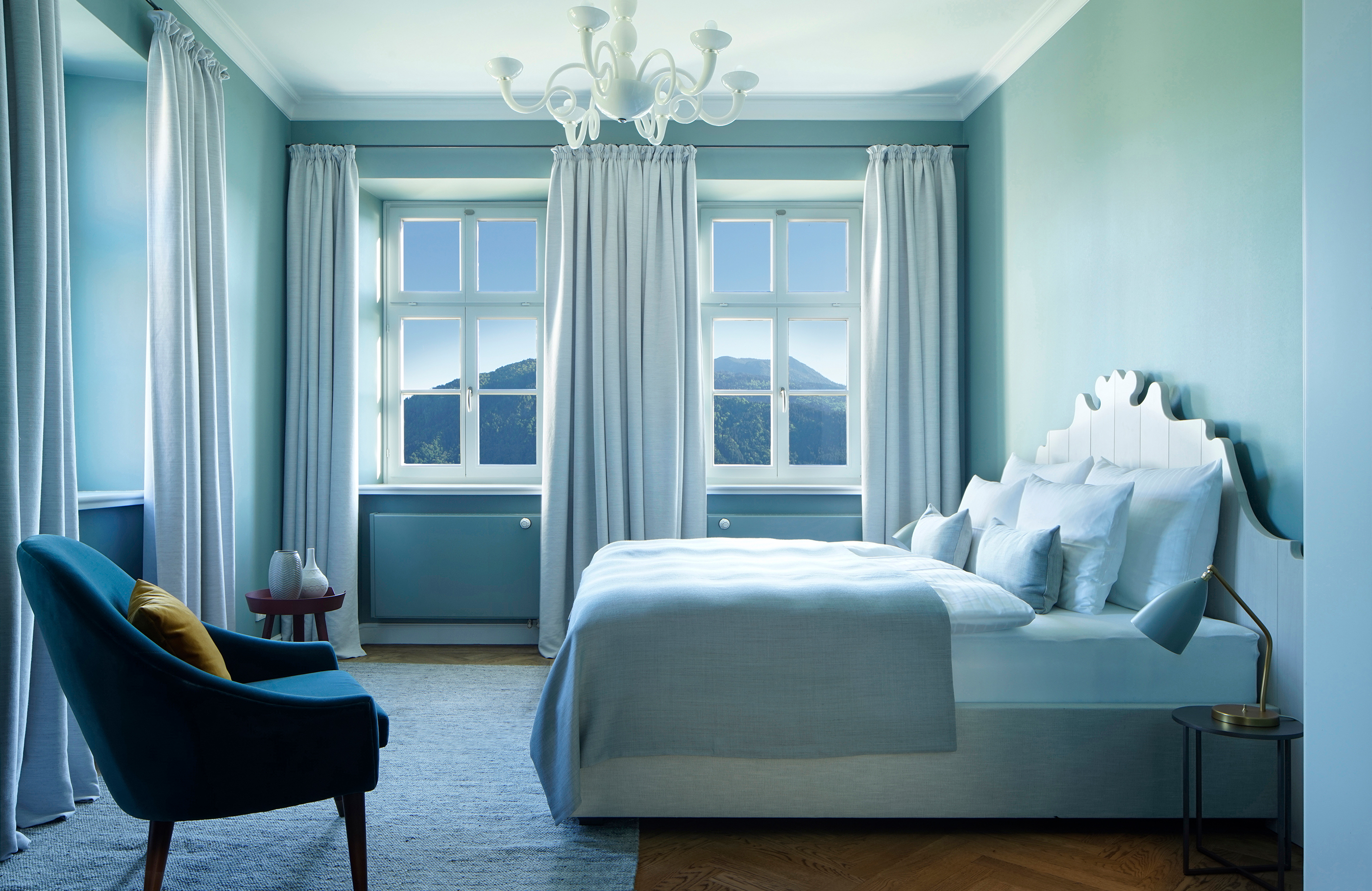Blick in eine Junior Suite im Hotel Das Tegernsee mit einem großen, weißen Doppelbett in der Raummitte, einem kleinen weißen Kronleuchter an der hohen Decke und mehreren Fenstern mit bodenlangen Vorhängen und Blick auf die Berge.
