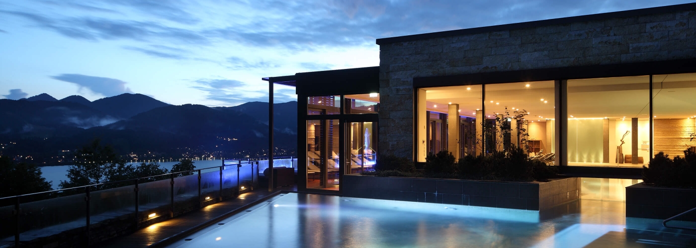 Aufnahme bei Nacht vom Hotel Das Tegernsee mit beleuchtetem Außenpool und dem Tegernsee mit den Bergen im Hintergrund. 