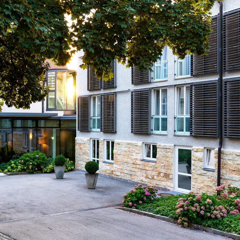 Sicht von außen auf die Herberge Quirin mit einer hellen Außenfassade und offenen, aufklappbaren Fensterläden aus Holz.