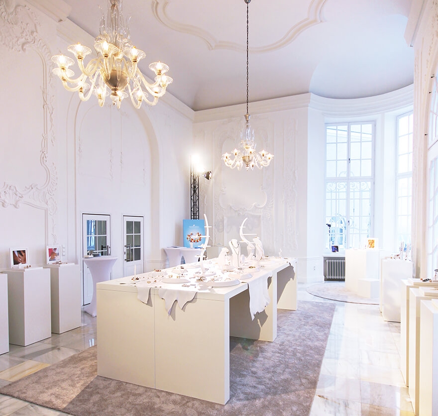 Aufnahme eines Raumes mit hohen Decken mit Barockelementen und Kronleuchtern, in dem gerade eine Ausstellung mit verschiedenen Schmuckstücken als Exponate statt findet.