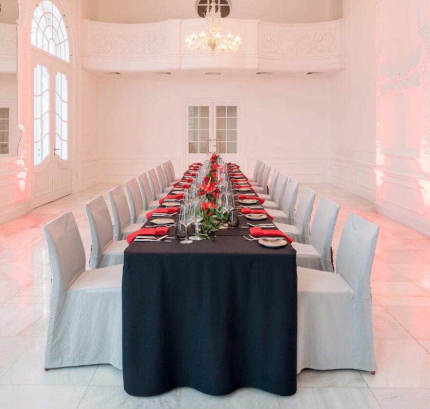 Aufnahme eines Veranstaltungsraums mit einer langen Tafel im Bildzentrum, die rot-schwarzen Farben eingedeckt ist und um die mehrere Stühle mit hellgrauen Stuhlhussen stehen.