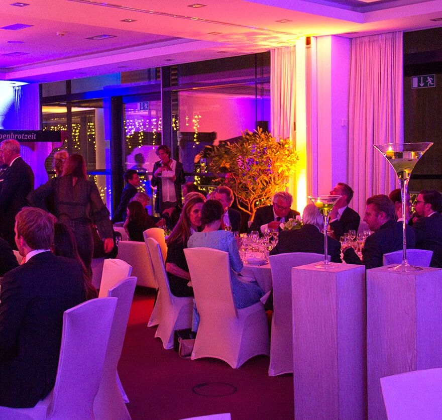 Blick in einen Veranstaltungsraum in dem sich eine Gesellschaft aus mehreren Personen befindet, die teilweise stehen und tweilweise an Rundtischen sitzen und sich in der Eventlocation am Tegernsee unterhalten.