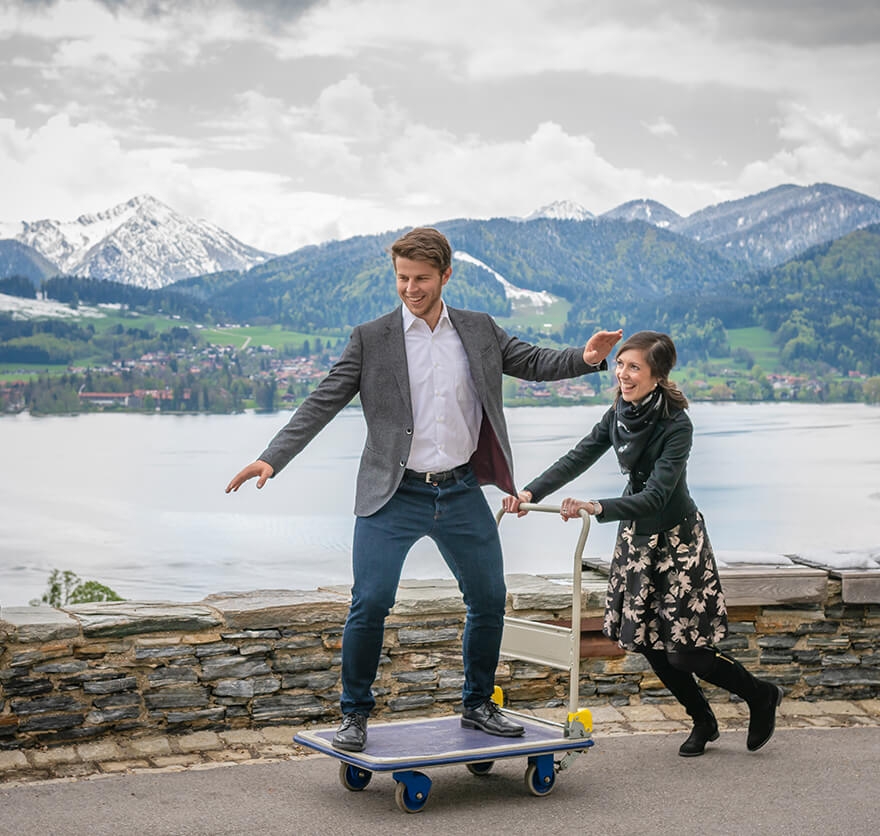 Eine Mitarbeiterin aus dem Hotel das Tegernsee schiebt lachend einen Kollegen, der stehend auf einem Lastenwagen balanciert, auf einem asphaltierten Weg direkt am Tegernsee mit den Bergen im Hintergrund voran.
