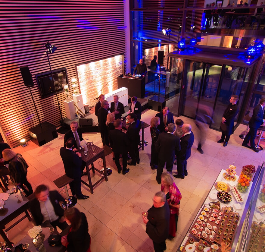 Blick von oben in den Empfangsbereich vom Hotel Das Tegernsee in dem mehrere Personen über den Raum verteilt teilweise an Stehtischen stehen, sich unterhalten und einige Getränke oder Canapés vom Buffet zu sich nehmen.
