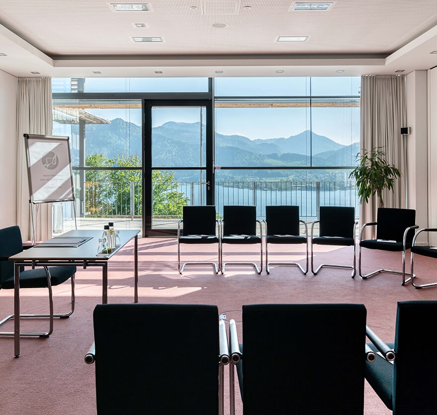 Aufnahme in einem Seminarraum im Hotel am Tegernsee mit mehreren in U-Form angeordneten Stühlen, im Zentrum ein weiterer Stuhl mit Tisch für den Referenten, ein Flipchart im Hintergrund und Blick durch die breite Fensterfront nach draußen auf den See und die Berge.