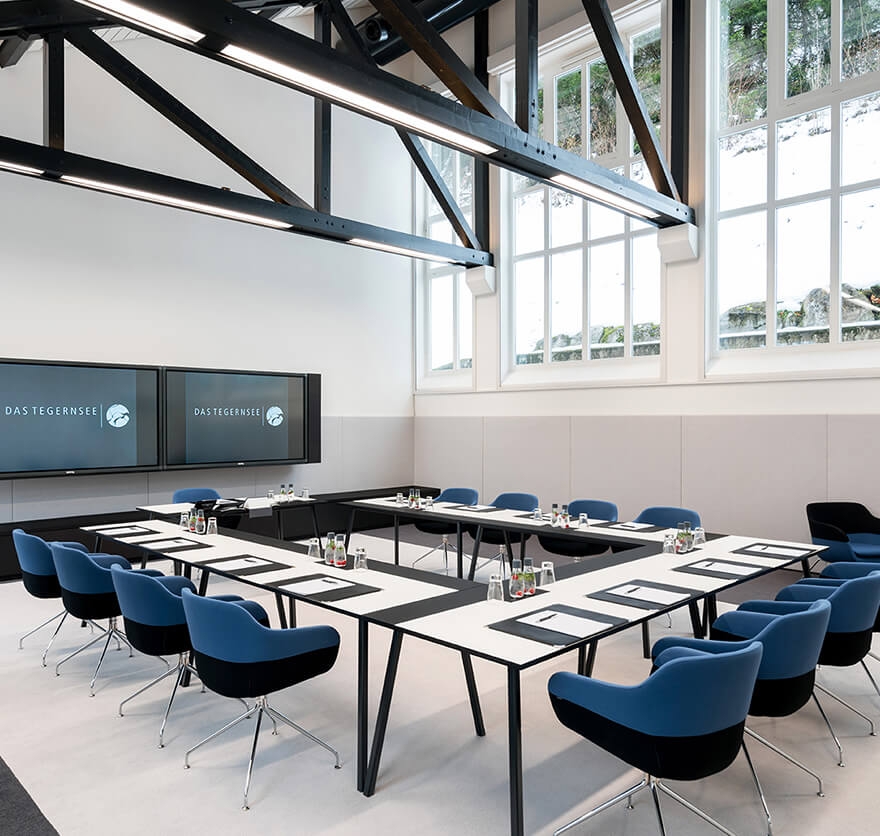 Aufnahme eines Tagungsraumes, in dem mehrere Tische und blau-schwarze Stühle in U-Form vor einem schwarzen Bildschirm, auf dem das Logo des Hotels Das Tegernsee angezeigt wird, aufgebaut sind.