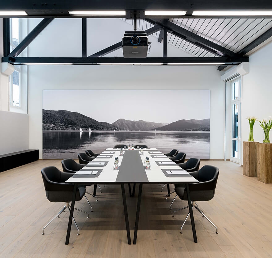 Blick in einen Tagungsraum im Tagungshotel Das Tegernsee mit einem langen, weißen Konferenztisch und schwarzen modernen Armlehnen-Stühlen im Bildzentrum und im Hintergrund ein großflächiges schwarz-weiß Bild vom Tegernsee und den Bergen.