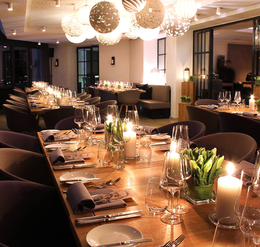 Aufnahme im Restaurant am Abend mit Blick auf mehrere eingedeckte Tische, auf denen angezündete Kerzen leuchten.