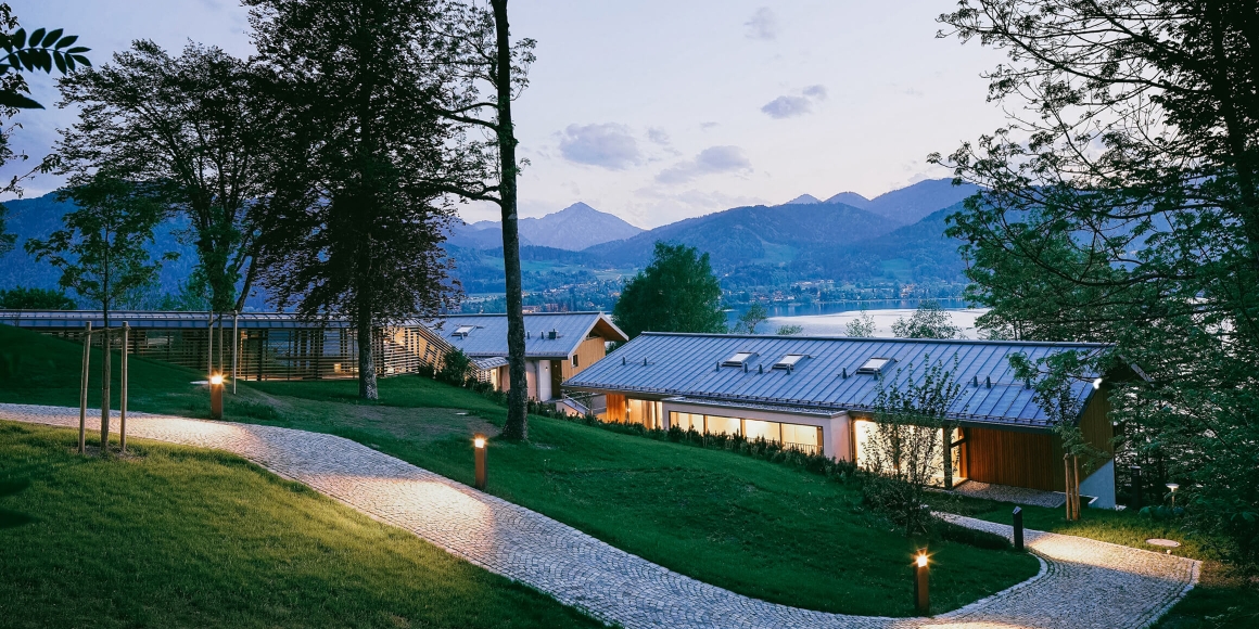 Blick von außen auf die Alpenchalets direkt am Tegernsee mit Blick auf die umliegenden Berge in der Abenddämmerung.