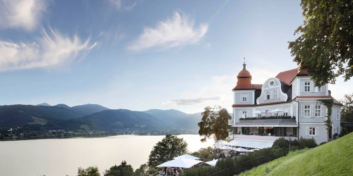 Außenaufnahme vom Hotel Das Tegernsee und dem Sengerschloss mit Blick auf den Tegernsee und die umliegenden Berge.