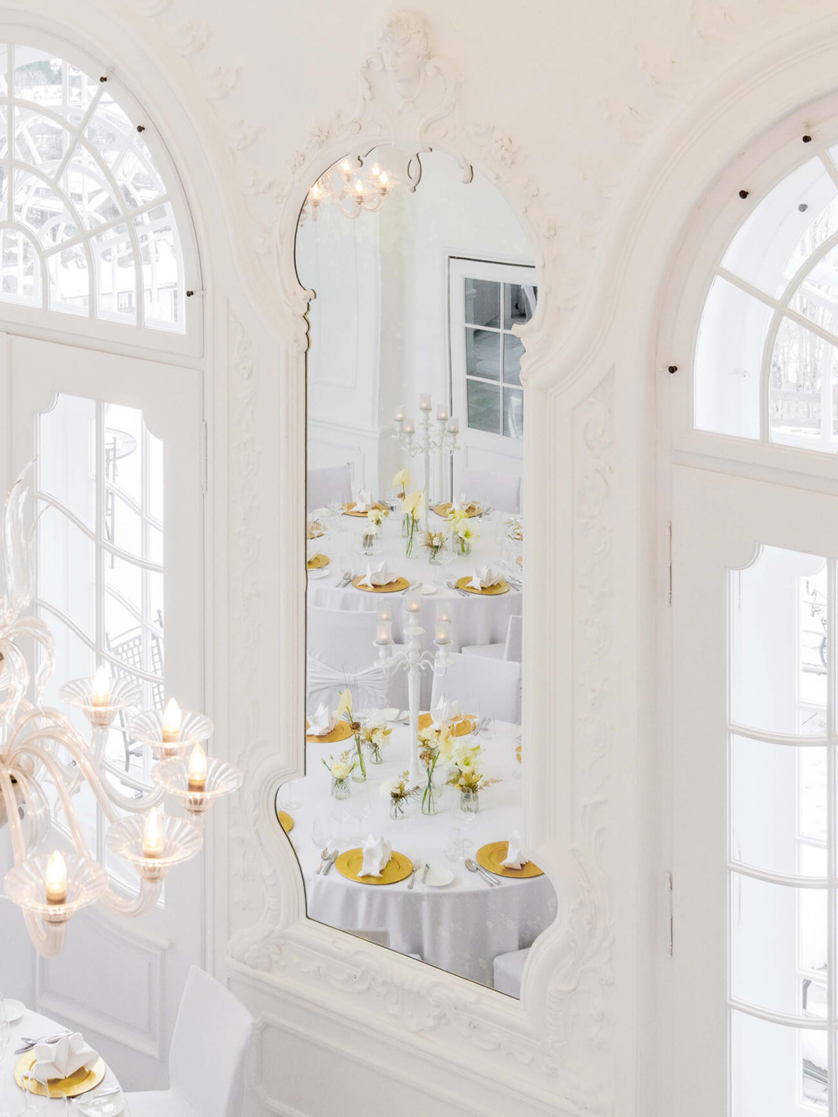 Detailaufnahme im Barocksaal mit einem großen Wandspiegel zwischen zwei hohen Bogenfenstern, in dem sich der festlich gedeckte Saal widerspiegelt.