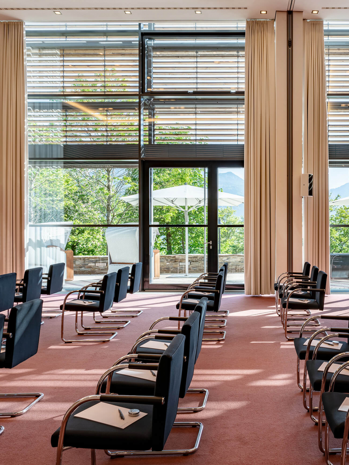 Aufnahme eines großen Tagungsraumes in dem mehrere Stühle in Reihen angeordnet sind mit einer langen Fensterwand mit direktem Zugang auf die Terrasse.