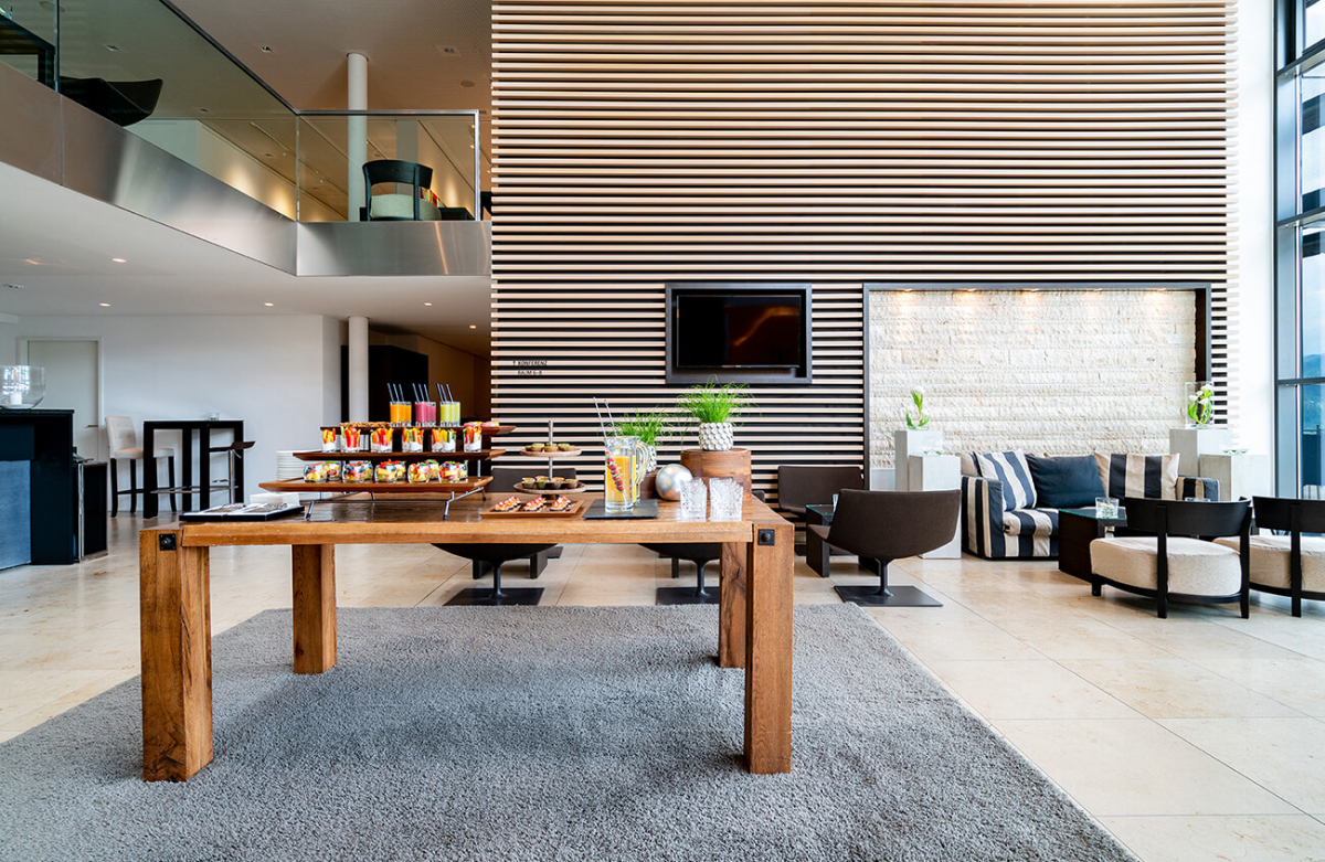 Aufnahme der weitläufigen Atrium Bar im Hotel Das Tegernsee mit einer Getränke- und Snack-Bar auf einem Holztisch und im Hintergrund moderne Sitzgelegenheiten wie Sofas und Sessel.